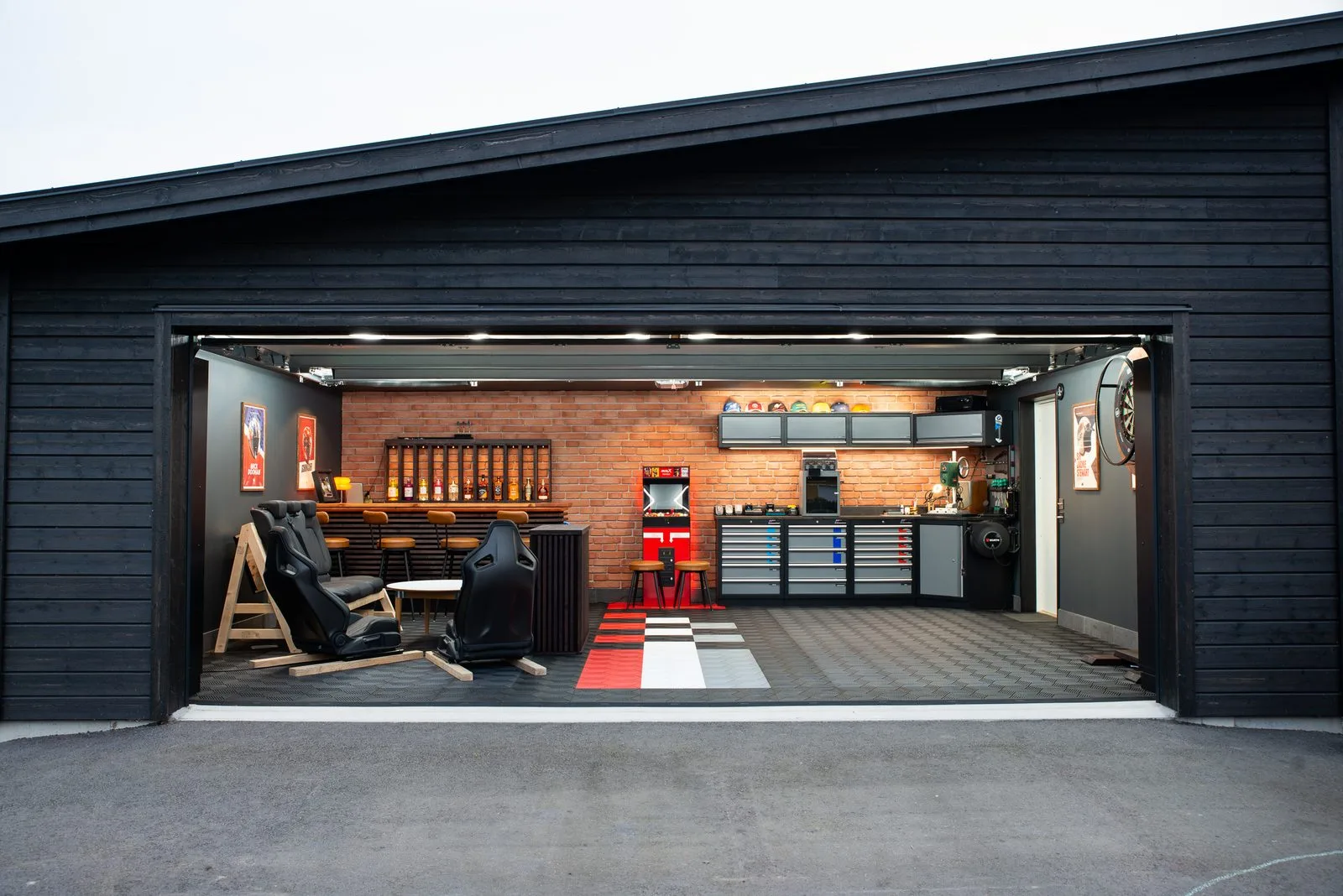 Svartbetsat garage med porten öppen, med bar, arkadspel och verkstadsskåp innanför