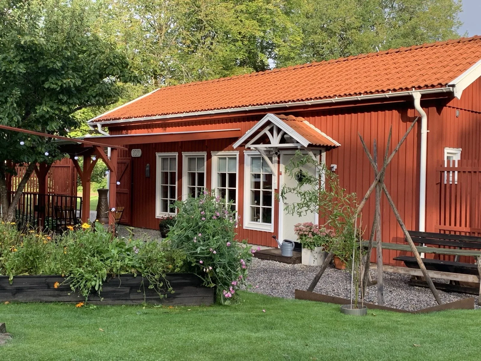 Faluröd byggnad med tegeltak, vita spröjsade fönster och vit dörr med skärmtak, omgiven av odlingslådor och gräsmatta